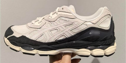 White sneaker with black accents, chunky sole, and mesh texture, held in a hand against a plain wall.